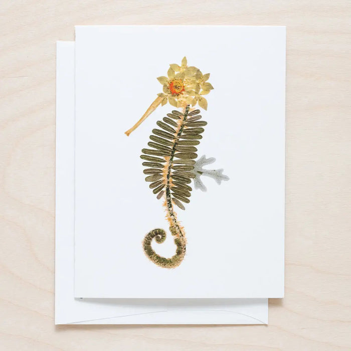 The PETAL PEOPLE PRESS - SEAHORSE CARD features a seahorse design made from pressed flowers and leaves, with green leaves for the body and yellow-brown flowers for the head and tail, set against a light wooden background.