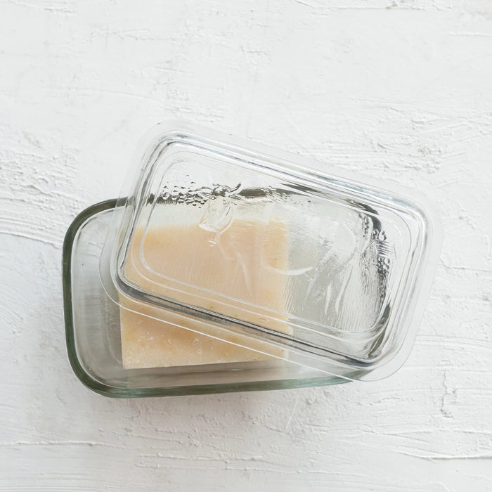 The CREATIVE COOP PRESSED GLASS BUTTER DISH WITH COW, featuring an embossed design and clear plastic lid, holds cheese or butter and looks perfect on a white textured surface for farmhouse kitchen decor.
