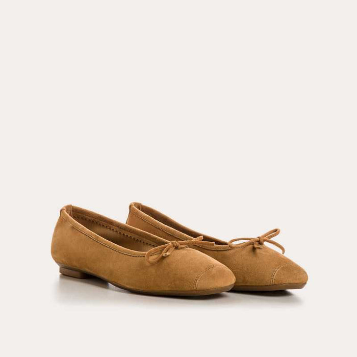 The REQINS - HARMONY SUEDE BALLERINA FLATS IN CAMEL feature a tan suede finish, a small bow on the toe, a low heel, and an elegant, simple design against a plain light background.