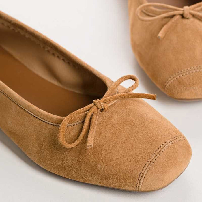 The REQINS - HARMONY SUEDE BALLERINA FLATS IN CAMEL feature a tan suede finish, a small bow on the toe, a low heel, and an elegant, simple design against a plain light background.