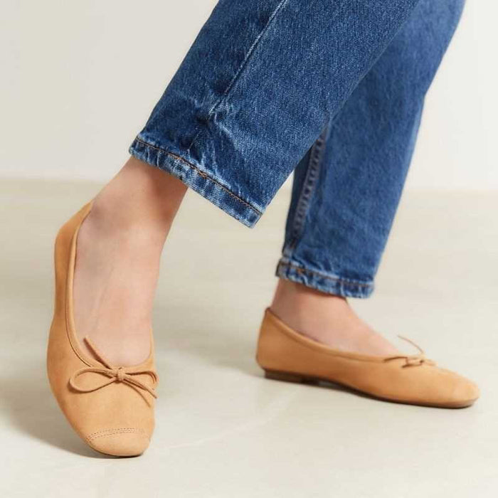 The REQINS - HARMONY SUEDE BALLERINA FLATS IN CAMEL feature a tan suede finish, a small bow on the toe, a low heel, and an elegant, simple design against a plain light background.