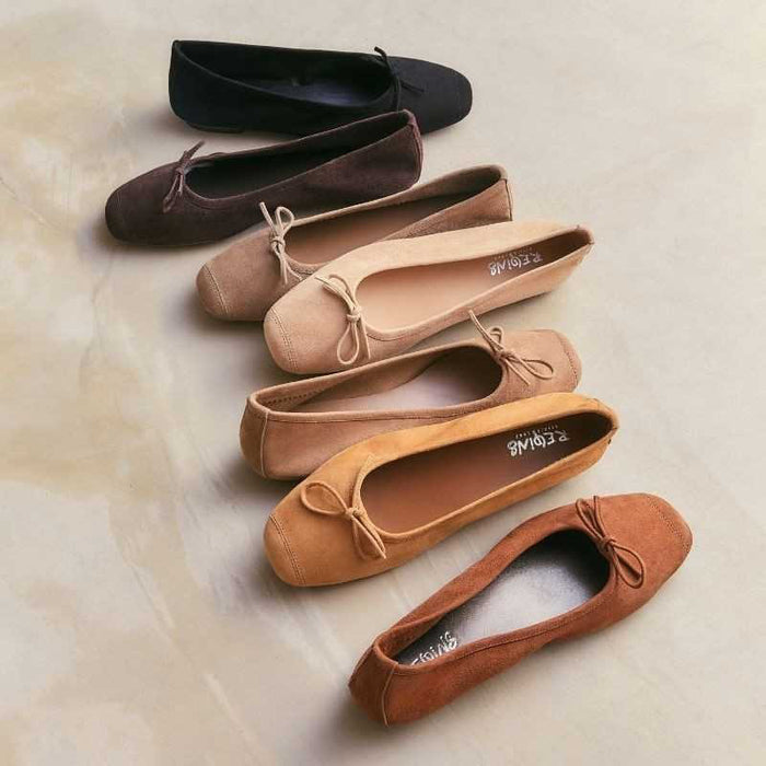 The REQINS - HARMONY SUEDE BALLERINA FLATS IN CAMEL feature a tan suede finish, a small bow on the toe, a low heel, and an elegant, simple design against a plain light background.