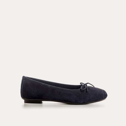 Someone stands on a light floor, one foot slightly raised, wearing REQINS - HARMONY suede ballerina flats in marine with a small bow, paired with blue jeans featuring frayed hems.