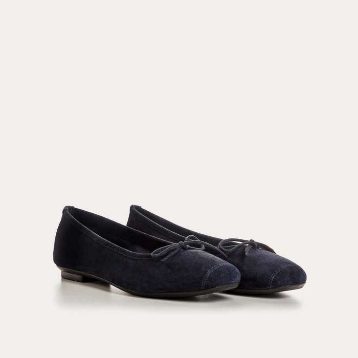 Someone stands on a light floor, one foot slightly raised, wearing REQINS - HARMONY suede ballerina flats in marine with a small bow, paired with blue jeans featuring frayed hems.