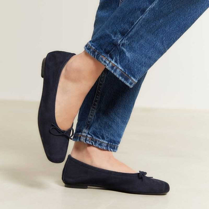 Someone stands on a light floor, one foot slightly raised, wearing REQINS - HARMONY suede ballerina flats in marine with a small bow, paired with blue jeans featuring frayed hems.