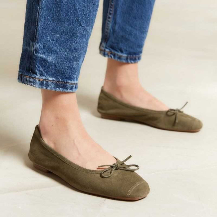 A person wearing olive green ballet flats with bows and cuffed blue jeans, standing on a light-colored floor.