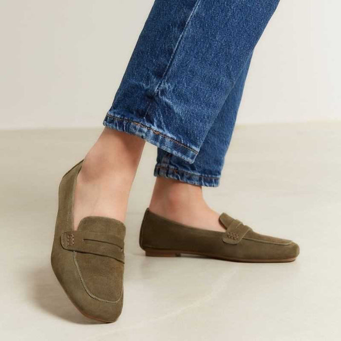 Person wearing olive green suede loafers and blue cuffed jeans, standing on a light-colored floor. The shoes have a classic penny loafer design with stitching details.
