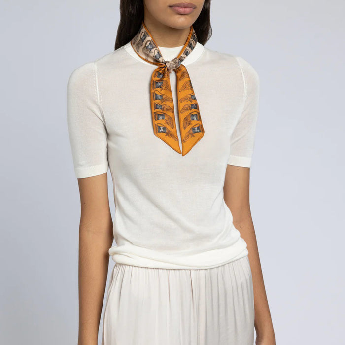 A woman wears a short-sleeved white top and pleated skirt, accessorized with the SABINA SAVAGE - THE TEMPLE OF PEGASUS SILK RIBBON from SABINA SAVAGE ENGLAND, standing against a plain light gray background.
