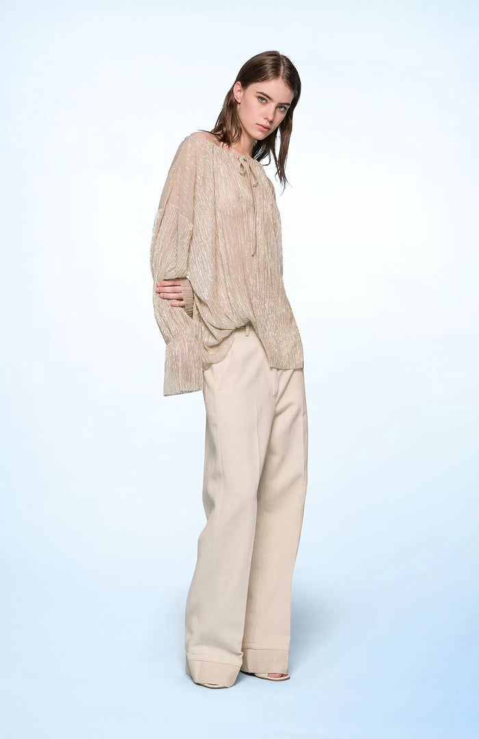 A woman with straight brown hair stands against a pale blue background, wearing the SEVENTY VENEZIA Lurex Jersey Blouse by SEVENTY with a gathered neckline and long sleeves, paired with wide-leg, high-waisted cream pants. She looks down in a relaxed pose.
