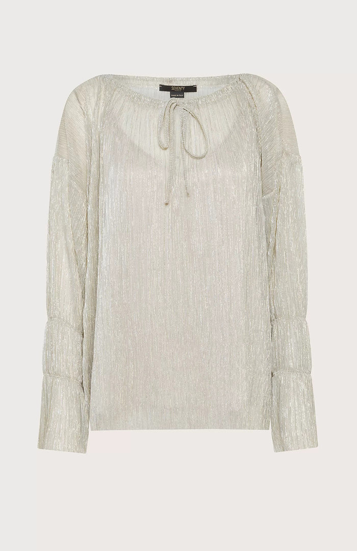 A woman with straight brown hair stands against a pale blue background, wearing the SEVENTY VENEZIA Lurex Jersey Blouse by SEVENTY with a gathered neckline and long sleeves, paired with wide-leg, high-waisted cream pants. She looks down in a relaxed pose.