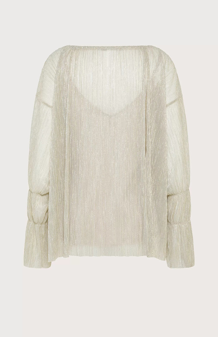 A woman with straight brown hair stands against a pale blue background, wearing the SEVENTY VENEZIA Lurex Jersey Blouse by SEVENTY with a gathered neckline and long sleeves, paired with wide-leg, high-waisted cream pants. She looks down in a relaxed pose.
