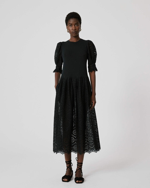 A woman stands against a white background wearing the SFIZIO - BRODERIE ANGLAISE MIDI DRESS by SFIZIO, featuring puffed sleeves and a lace skirt, paired with black strappy sandals.