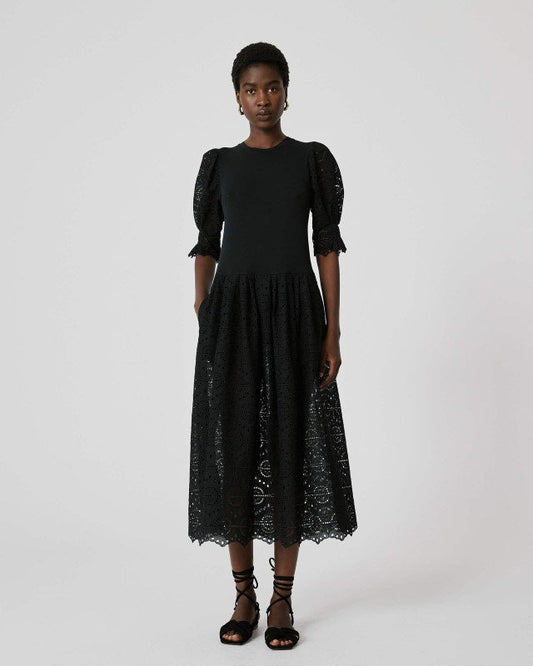A woman stands against a white background wearing the SFIZIO - BRODERIE ANGLAISE MIDI DRESS by SFIZIO, featuring puffed sleeves and a lace skirt, paired with black strappy sandals.