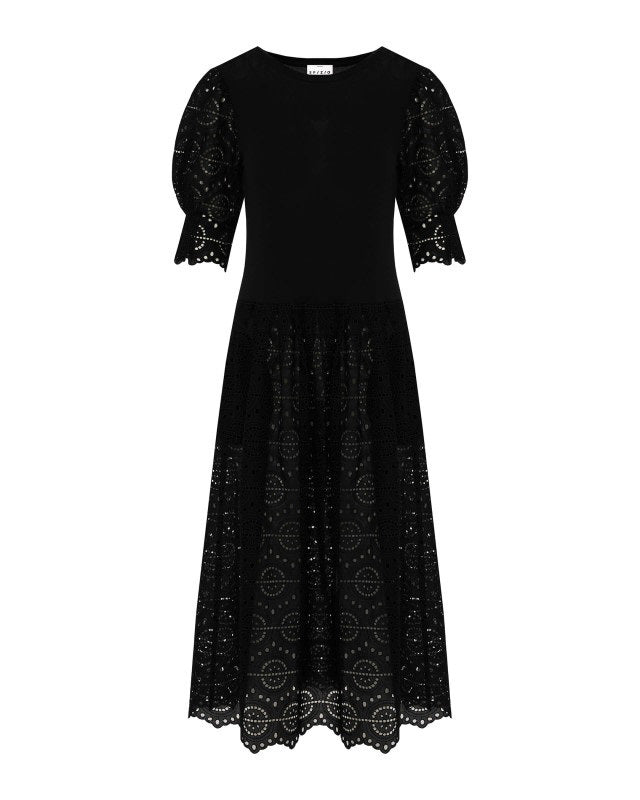 A woman stands against a white background wearing the SFIZIO - BRODERIE ANGLAISE MIDI DRESS by SFIZIO, featuring puffed sleeves and a lace skirt, paired with black strappy sandals.