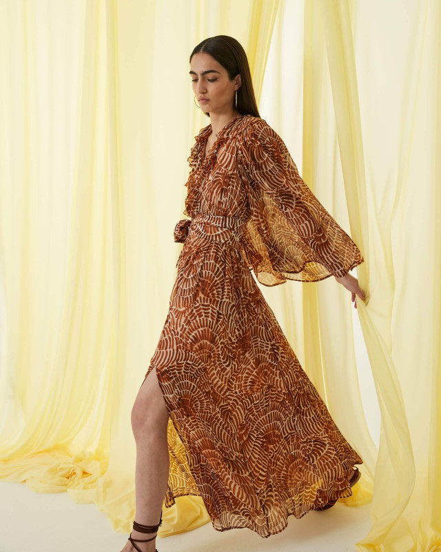 The SFIZIO - RUST PRINT RUFFLED DRESS by SFIZIO is a long-sleeved, floor-length Italian dress in airy viscose creponne with a cinched waist, ruffled neckline, and abstract brown/beige print on lightweight, semi-sheer fabric.