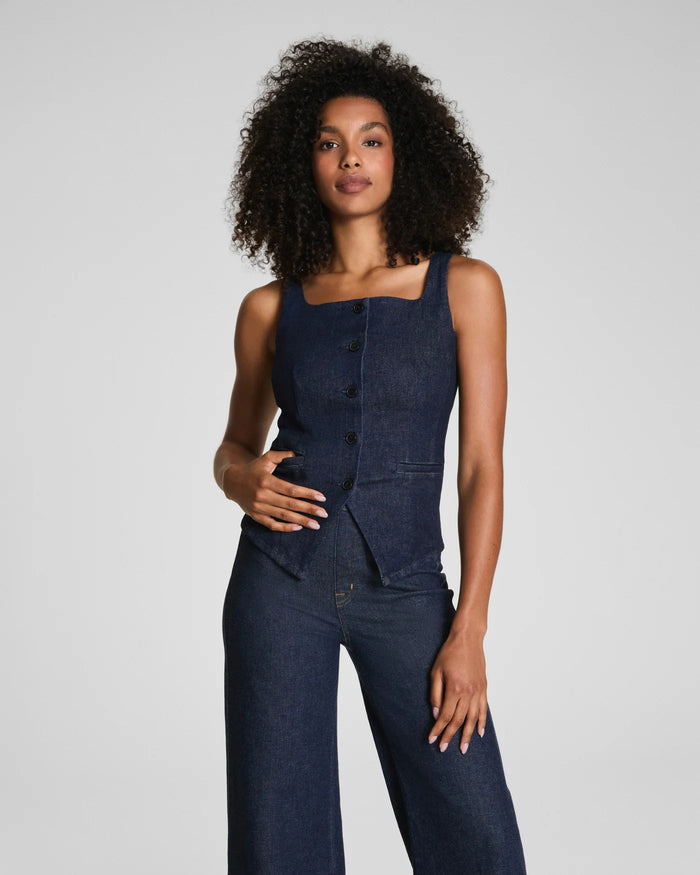 A person with curly hair faces away, showcasing the SPANX - DENIM TAILORED VEST and matching wide-leg jeans in dark blue cotton blend against a plain light gray background.