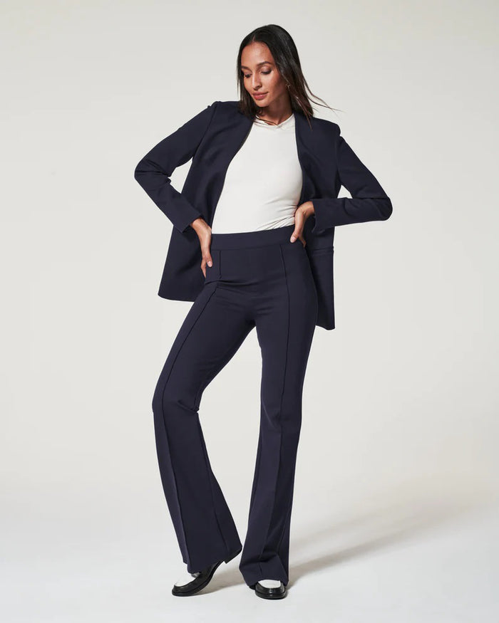 A woman models the SPANX Perfect Pant High Rise Flare in navy with a matching blazer, white fitted top, and black-and-white shoes, highlighting the sleek pull-on design from SPANX against a plain light background.