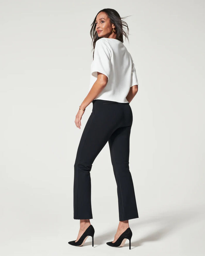 A woman stands confidently with hands on hips, wearing a white short-sleeve top, black SPANX - PERFECT PANT KICK FLARE from SPANX, and black heels against a plain light background.