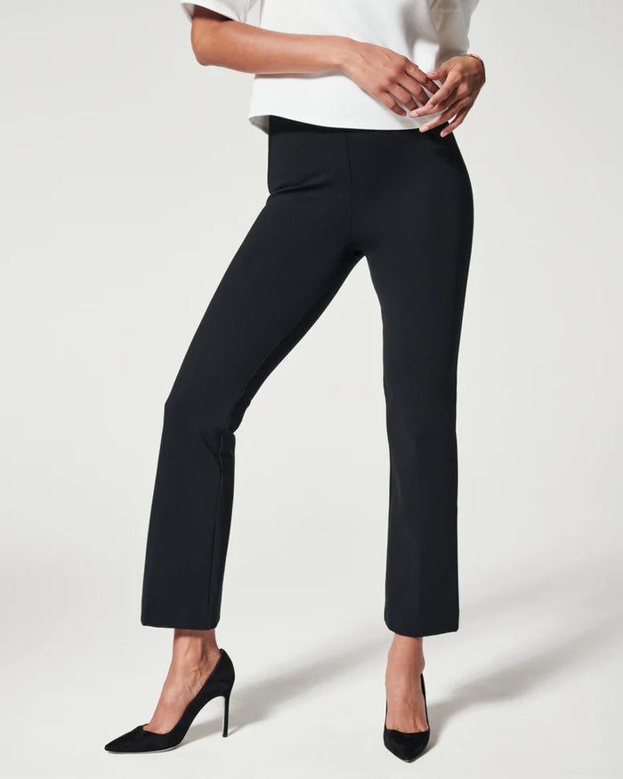 A woman stands confidently with hands on hips, wearing a white short-sleeve top, black SPANX - PERFECT PANT KICK FLARE from SPANX, and black heels against a plain light background.