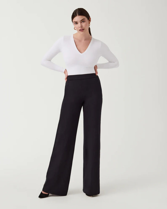 A woman stands against a plain background wearing the SPANX Perfect Pant Wide Leg in black—crafted from premium ponte fabric—paired with a white long-sleeve V-neck top and black heeled shoes.