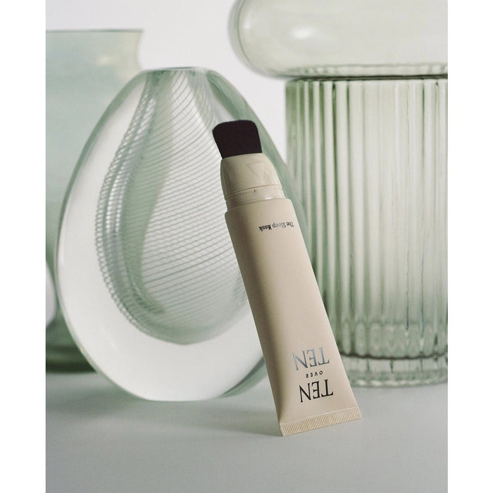 A beige tube labeled "TEN OVER TEN - THE SLEEP MASK" is displayed horizontally in front of textured glass vases on a soft, light background—an elegant way to showcase this luxurious hand cream from TEN OVER TEN.