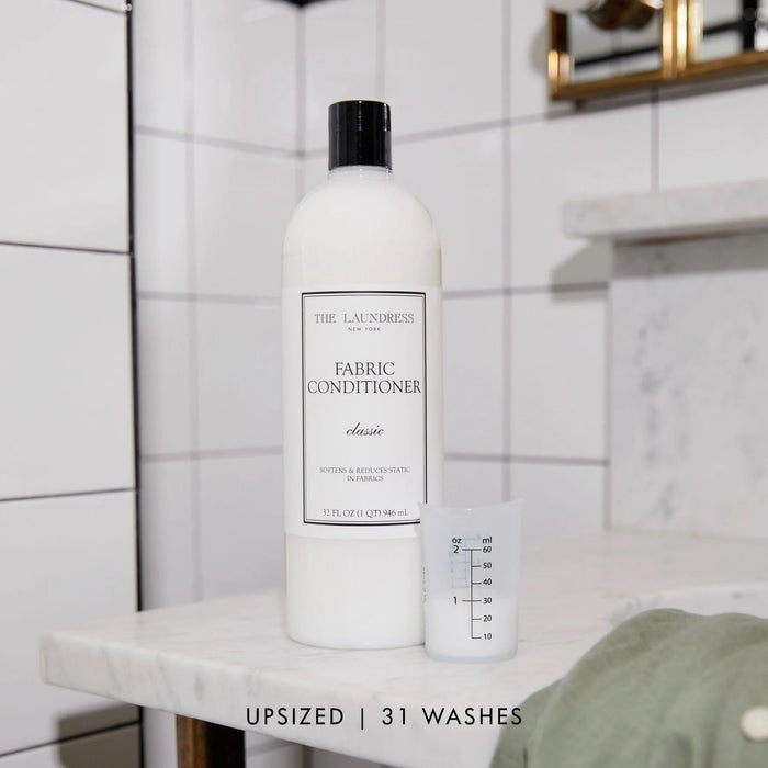 THE LAUNDRESS - FABRIC CONDITIONER 32OZ is a plant-derived fabric softener that helps reduce static. It comes in a clear plastic bottle with a black cap and contains 32 fl oz (946 ml).