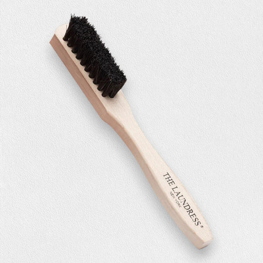 THE LAUNDRESS - STAIN BRUSH features black bristles and "THE LAUNDRESS NEW YORK" on the wooden handle; perfect for fabric stain removal and shown on a light gray background.