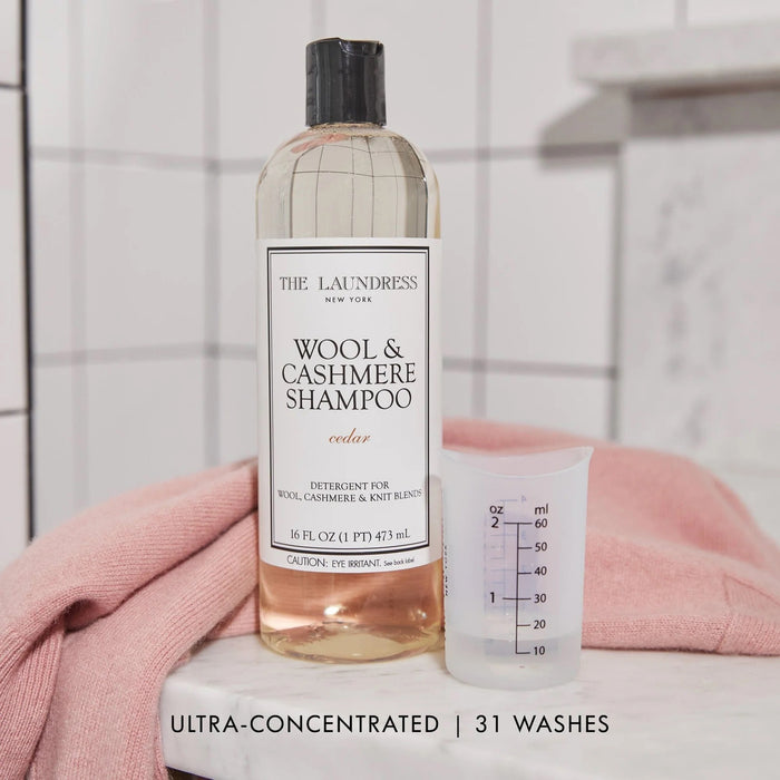 A 16oz clear plastic bottle of THE LAUNDRESS WOOL AND CASHMERE SHAMPOO, cedar-scented, with a white label and black text. This enzyme-free detergent is perfect for gently cleaning wool and knit blends.