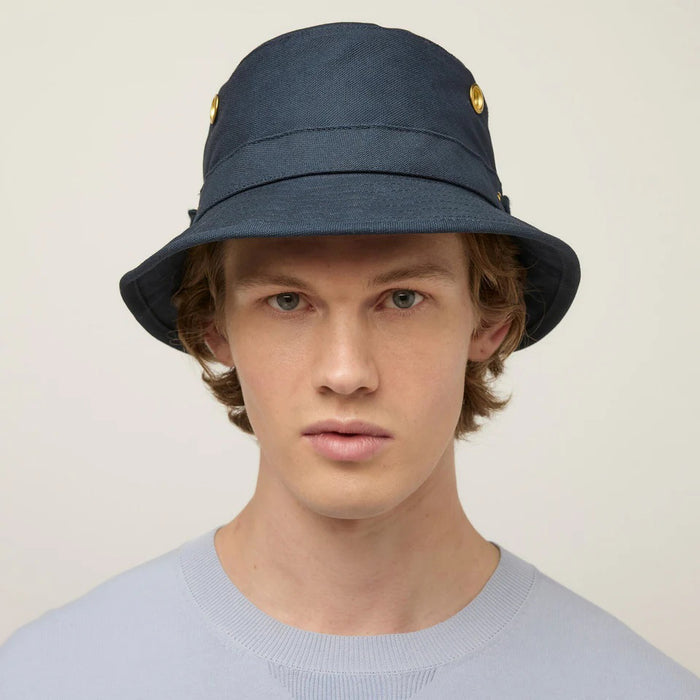 A young person with fair skin and light brown hair wears the TILLEY - ICONIC T1 BUCKET HAT IN DARK NAVY and a light gray crew-neck shirt, looking directly at the camera against a plain, light background.