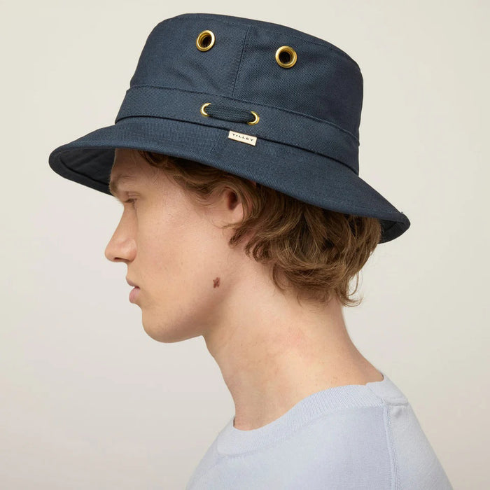 A young person with fair skin and light brown hair wears the TILLEY - ICONIC T1 BUCKET HAT IN DARK NAVY and a light gray crew-neck shirt, looking directly at the camera against a plain, light background.