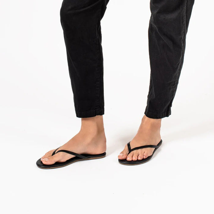 A pair of TKEES - LILY SLINER SLIPPER SABLE by TKEES INC, featuring black flip-flop sandals with thin straps and “TKEES” in gold on the cushioned insole, shown on a white background.