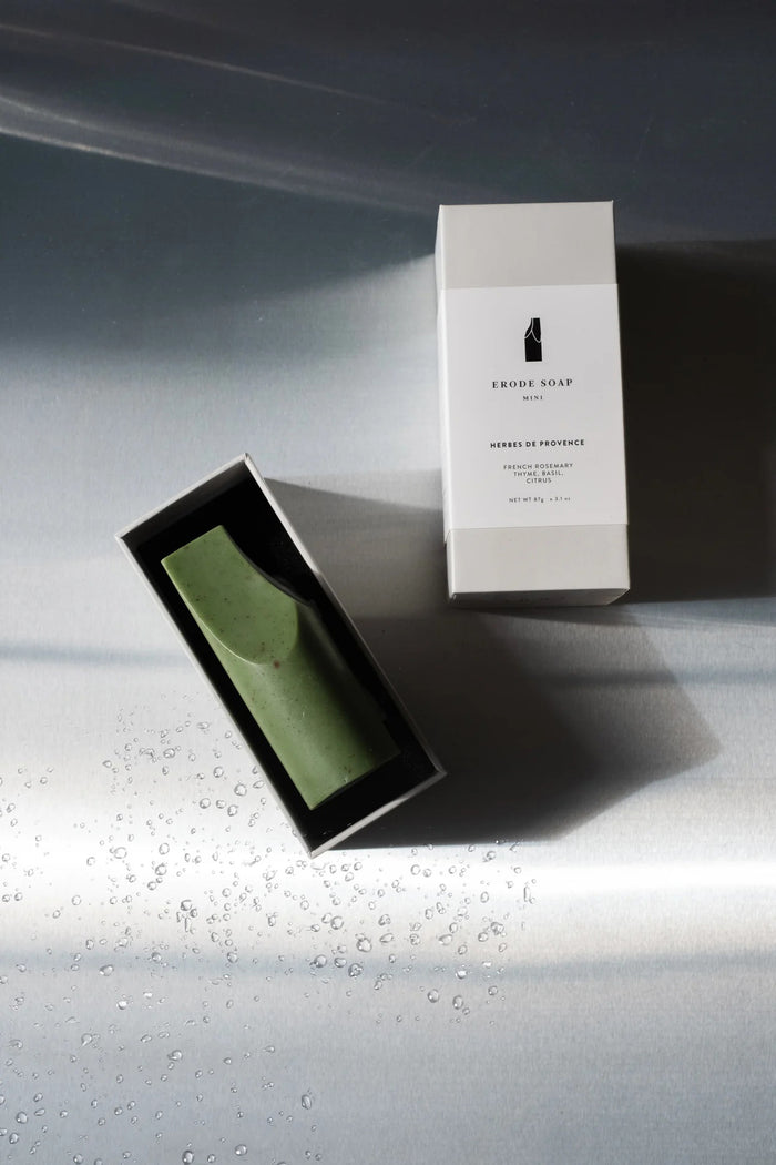 The UME - HERBES DE PROVENCE ERODE MINI SOAP by UME STUDIO is a green, smooth, abstract soap with a tall cylindrical base, slanted flat top, and curved cut on one side, shown against a light gray background.