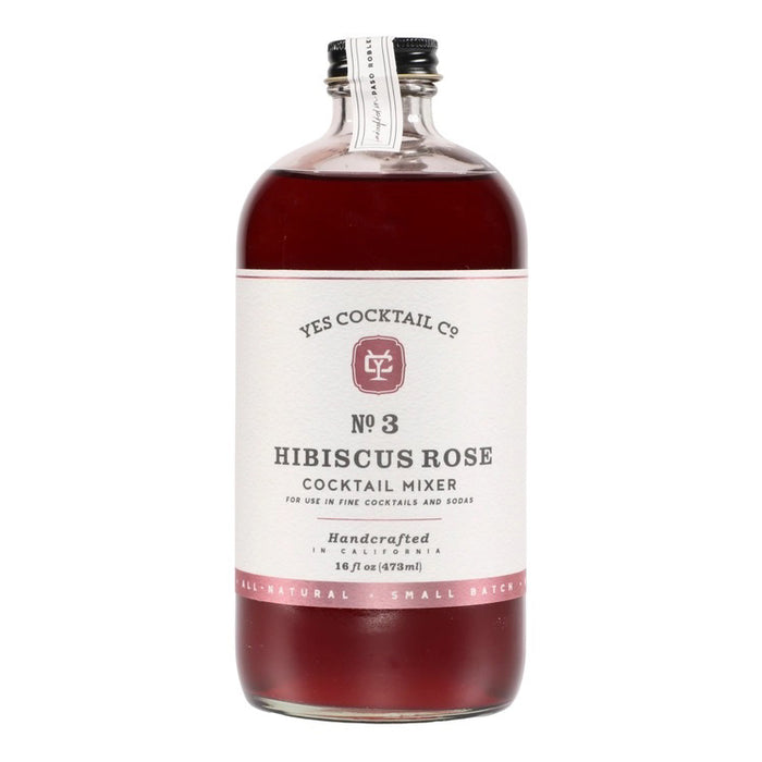 A transparent glass bottle holds a rich, dark red liquid labeled "YES COCKTAIL CO - Hibiscus Rose Cocktail Mixer." Handcrafted in California and available in a 16 fl oz (473ml) size, the white-and-red-toned label indicates its perfect suitability for creating cocktails or an exotic floral spritzer.